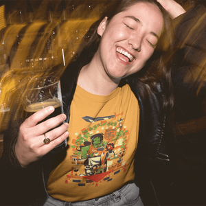 Person wearing a Legends and Lattes T-shirt with a graphic design, holding a glass of dark beer, with a blurred background.